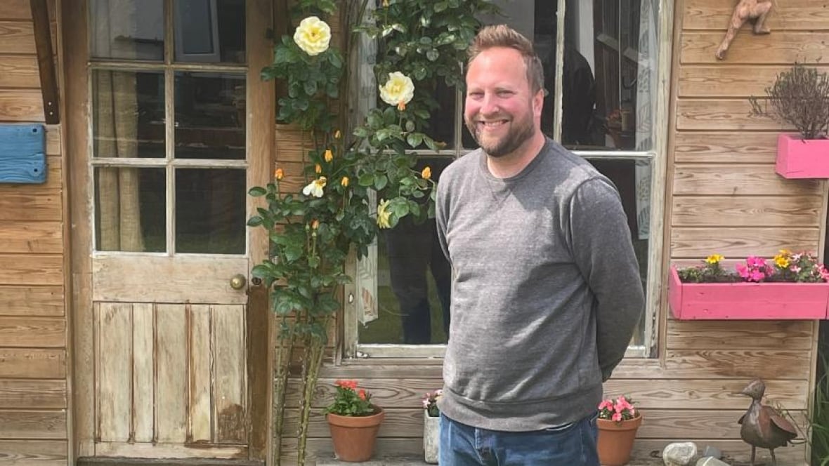 A man in a sweat stands in front of a garden shed smiling. 
