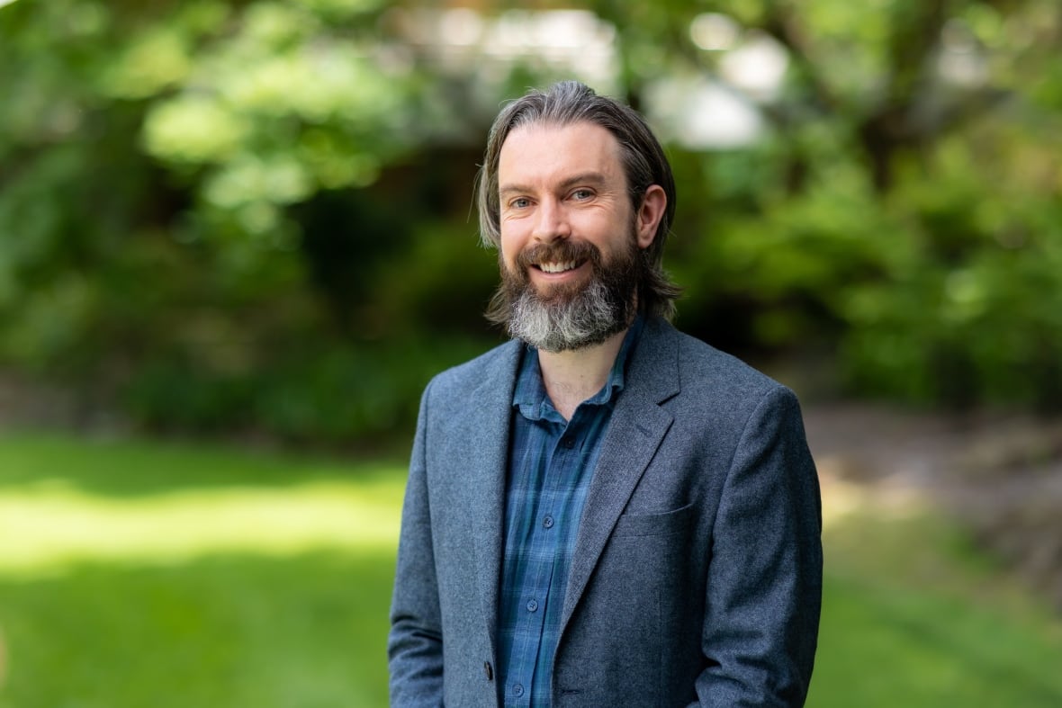 A man wearing a suit and with a beard looks into a camera lens while in a garden. 
