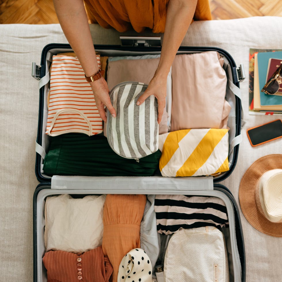 shot from above of an anonymous woman packing things in her suitcase on the bed she is holding and putting a grey and white stripped cosmetics bag in there is her mobile phone, books and sunglasses on the bed