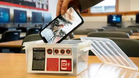 PA Media Image shows a mobile phone being put inside a clear plastic box in a classroom.